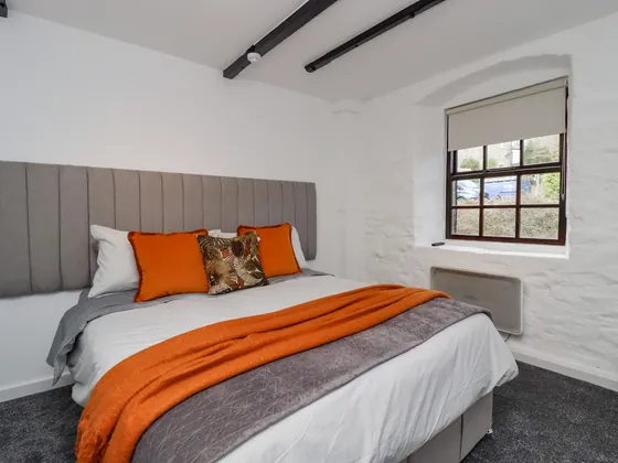 A double bed with grey bedding and orange cushions under a small window